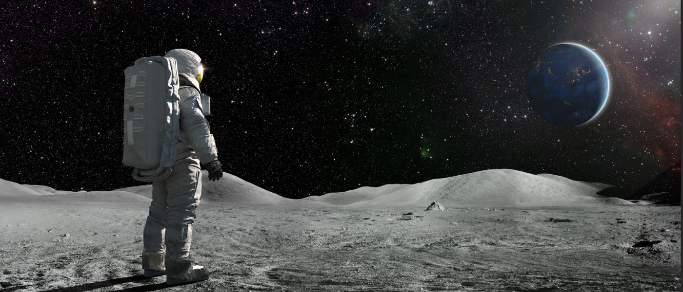 A lone astronaut standing facing away from the camera dressed in full space suit with backpack, stands still looking towards a distant planet Earth. The sun illuminates a side of Earth and hundreds of stars are visible in deep space.
