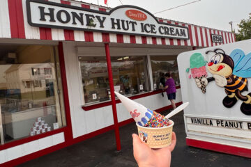 Photo of the Honey Hut building and sign with a hand holding ice cream in a cup in front of it