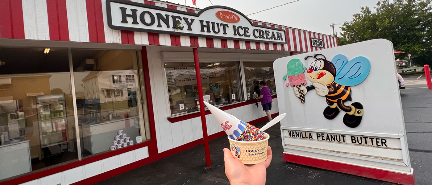 Photo of the Honey Hut building and sign with a hand holding ice cream in a cup in front of it