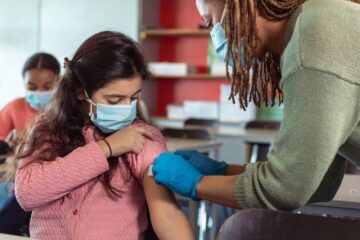 School nurse placing bandage on a female elementary age student's arm after administering a vaccine. The medical professional and student are both wearing protective face masks.