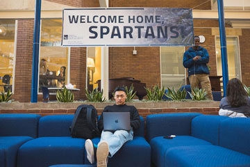 Photo of a student sitting on a couch in Thwing Center with their laptop and a banner that says "Welcome Home Spartans" behind them