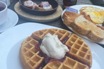 Close up of breakfast foods, including a Belgian waffle, cup of fruit, eggs, potatoes, toast and cinnamon rolls).