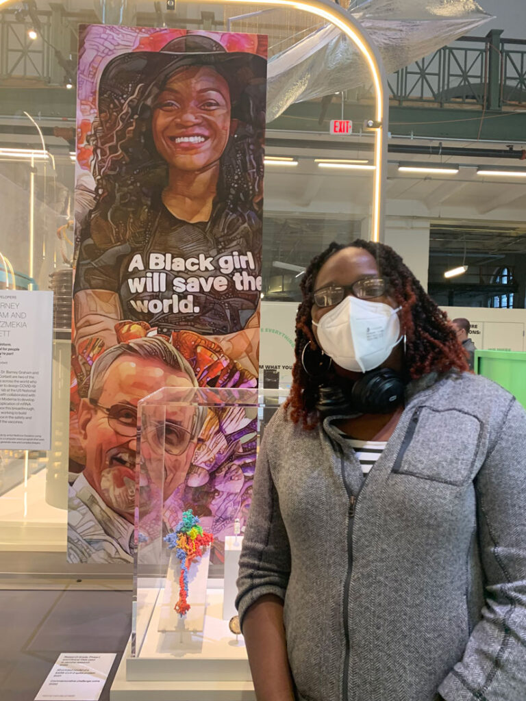 Photo of Olubukola Abiona in front of an exhibit at the Smithsonian that has a banner with an illustration of a Black woman wearing a shirt that says "A Black girl will save the world"