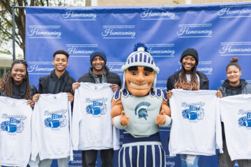 Photo of five students holding up homecoming T-shirts as they pose for a photo with Spartie