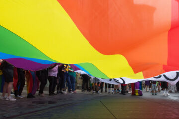 Photo taken from below a rainbow flag spread out and held by many people at a pride parade