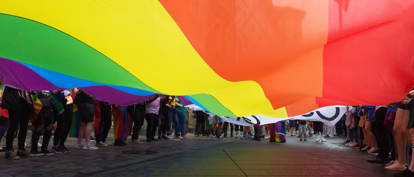 Photo taken from below a rainbow flag spread out and held by many people at a pride parade