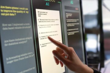 Close-up stock photograph showing a touchscreen monitor being used in an open plan office. A woman’s hand is asking an AI chatbot pre-typed questions & the Artificial Intelligence website is answering.