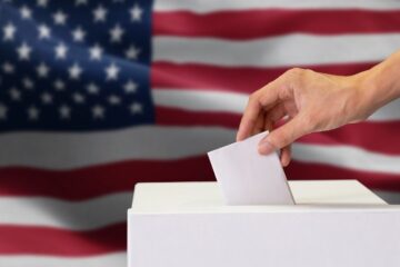 Close-up of man casting and inserting a vote and choosing and making a decision what he wants in polling box with United States flag blended in background. Courtesy of Getty Images.