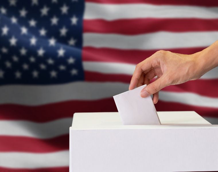 Close-up of man casting and inserting a vote and choosing and making a decision what he wants in polling box with United States flag blended in background. Courtesy of Getty Images.