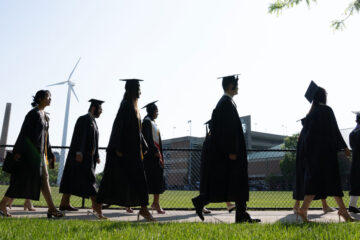 Photo of Class of 2023 graduates in their caps and gowns walking past Van Horn Field to the Veale Center
