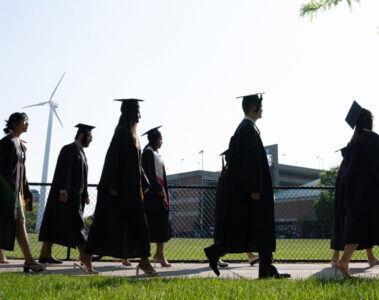Photo of Class of 2023 graduates in their caps and gowns walking past Van Horn Field to the Veale Center