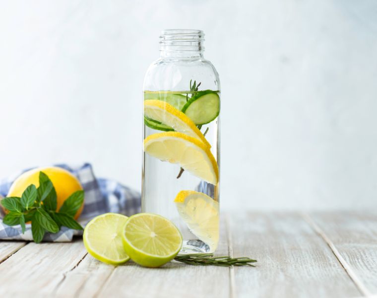 Bottle of infused water on white wood with a slice of lemon , cucumber and rosemary leaf in it.Courtesy of Getty Images.
