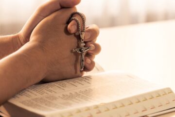 Close up of folded hands praying on holy bible book while holding a pendant crucifix.