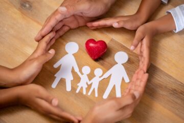 Close up hands of father, mother and children protecting family paper cutout with red heart.Photography by Getty Images.