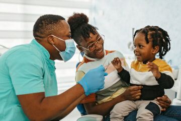 Photo of a dentist examining a smiling child's teeth while he sits in his mom's lap