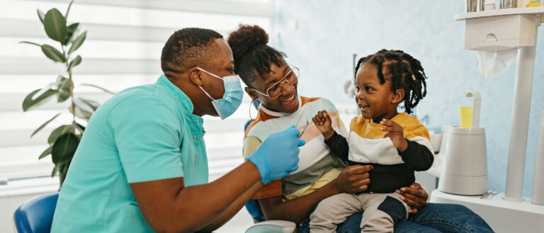 Photo of a dentist examining a smiling child&#039;s teeth while he sits in his mom&#039;s lap