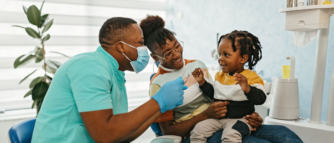 Photo of a dentist examining a smiling child's teeth while he sits in his mom's lap