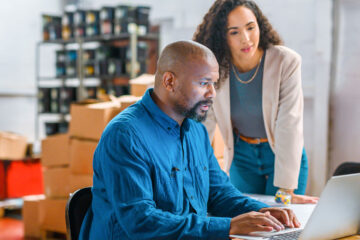 Photo of two colleagues working together at a laptop with boxes behind them in an office