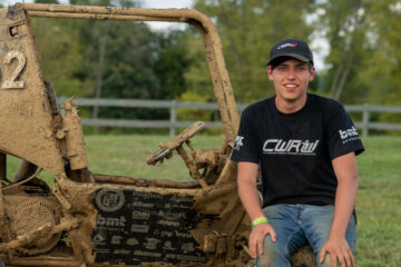 Photo of Daniel Clare sitting on the muddy CWRU Motorsports Baja SAE car