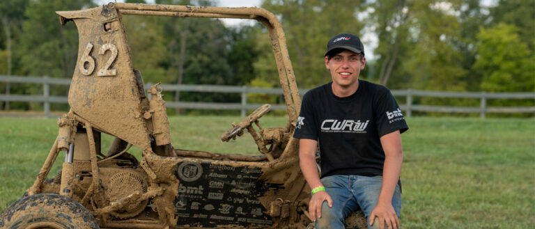 Photo of Daniel Clare sitting on the muddy CWRU Motorsports Baja SAE car