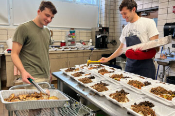 Photo of Peter Gittings and Jacob Boodjeh filling to-go boxes with food on opposite sides of a worktop in the kitchen