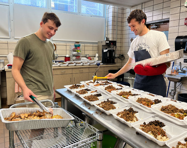 Photo of Peter Gittings and Jacob Boodjeh filling to-go boxes with food on opposite sides of a worktop in the kitchen