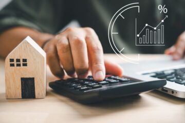 Close up of an individual typing on a calculator next to a miniature wooden house. Courtesy of Getty Images.