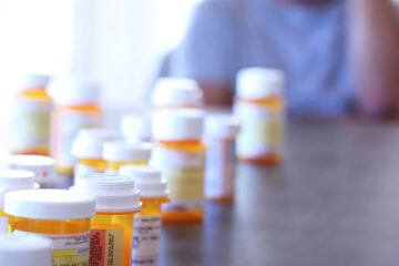 Photo of prescription pill bottles lined up on a table with a man in distress blurred in the background