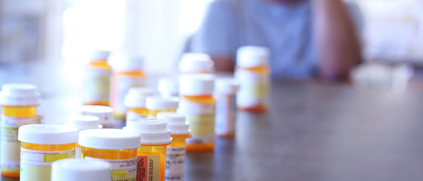 Photo of prescription pill bottles lined up on a table with a man in distress blurred in the background