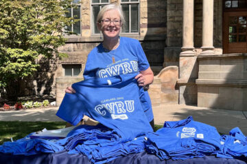 Photo of CWRU staff member Cindy Young wearing a CWRU T-shirt while holding up another and standing next to a table with a pile of more