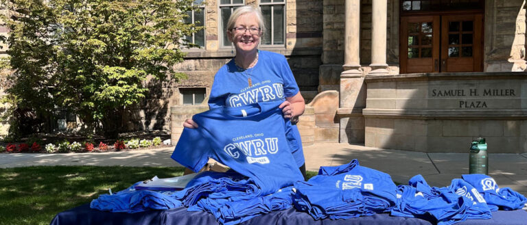 Photo of CWRU staff member Cindy Young wearing a CWRU T-shirt while holding up another and standing next to a table with a pile of more