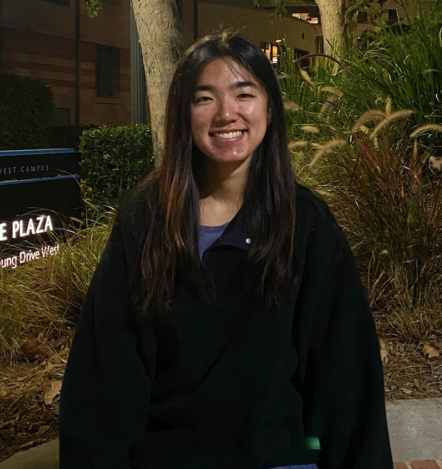 A student stands in a long sleeve black quarter zip outside in front of trees and foliage, smiling. 