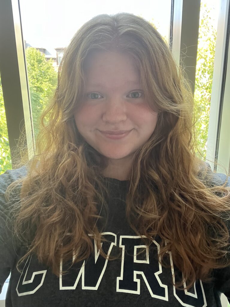 A woman student with long red hair in a CWRU shirt stands in front of a window, smiling. 