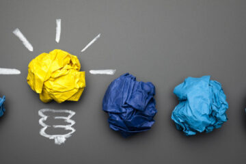 Line of five crumpled balls of paper, with one in yellow and a drawing around it to make it look like a light bulb