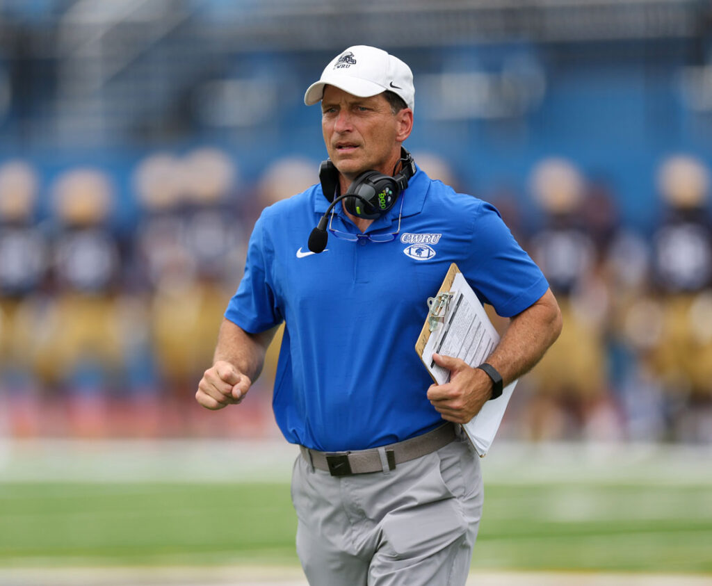 Photo of Greg Debeljak holding a clipboard and watching from the sidelines of a CWRU football game