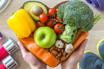 Photo of someone holding a heart-shaped dish full of nutritious foods with water bottles, tennis shoes and weights on the floor