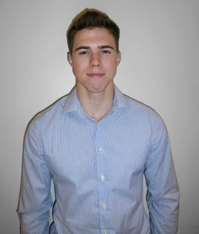 A male student stands in front of a white wall with a blue button up shirt on. 