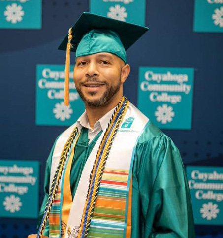 A male student stands in a green graduation cap and gown in front of a "Cuyahoga Community College" backdrop. 