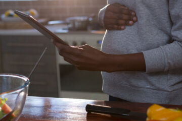 Photo of a pregnant woman looking at a tablet with fresh food on the counter in front of her