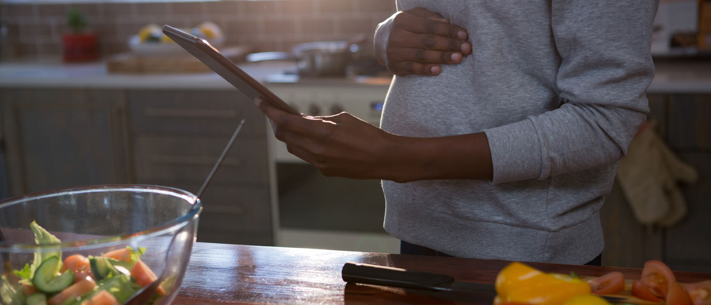 Photo of a pregnant woman looking at a tablet with fresh food on the counter in front of her